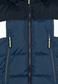 Doudoune marine avec une section supérieure noire horizontale, deux poches latérales zippées et des tirettes de zip vert clair, présentant une texture matelassée.