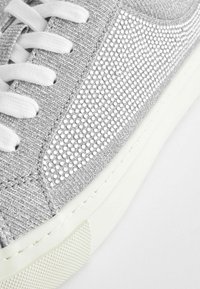 Grå sneaker med en texturerad ovandel som har metalliska detaljer, vita snören och en gummisula i vitt med ett mönstrat grepp.