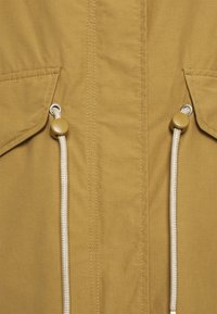 Beżowa kurtka z fakturowanej tkaniny, wyposażona w dwa regulowane sznurki z beżowymi stoperami oraz widoczne przeszycia z przodu.