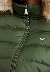 Gros plan d'une veste matelassée verte avec capuche bordée de fourrure, fermeture éclair centrale et petit logo rouge, blanc et bleu sur la poitrine.