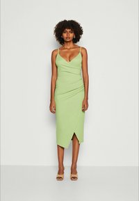 Vestido verde ajustado con tirantes de espagueti ajustables, diseño envolvente en la parte delantera, hasta la rodilla y con textura suave. Llevado con sandalias beige.