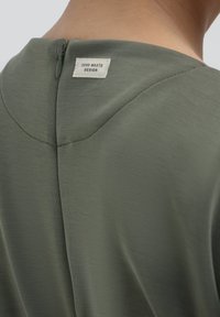 Indumento verde oliva con scollo rotondo, dotato di una chiusura con zip sul retro e un'etichetta "Zero Waste Design" vicino al collo. Texture liscia.