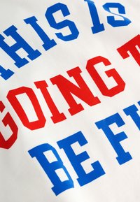 T-shirt in tessuto bianco con lettere audaci e a blocchi. Testo rosso con la scritta "GOING TO" e blu con "BE FUN", con una texture liscia di cotone.