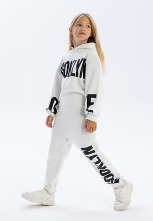 Sweatshirt à capuche blanche et joggers assortis avec un texte noir audacieux. Tissu lisse, coupe décontractée et poignets élastiques aux chevilles.