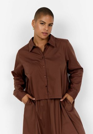 Selvsikker person med kort hår, der bærer guldringe og en løs, brunt knapbluse med matchende bukser, hænderne i lommerne, neutral baggrund.