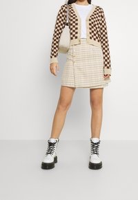 Braun und beige kariertes Strickjacke über einem weißen Hemd, kombiniert mit einem beigen karierten Rock und weißen Schnürstiefeletten.