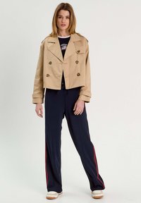 Chaqueta beige de corte recto con doble botonadura y grandes botones, combinada con pantalones navy con rayas rojas en los laterales y zapatillas blancas. Textura de tela suave.