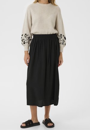 Femme portant un pull beige avec des manches florales noires, une jupe midi noire taille haute et des sandales noires ouvertes à l’avant sur fond blanc.