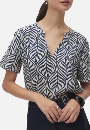 Mujer con blusa azul marino y blanca con estampado de hojas y escote en V, pantalones oscuros con cinturón, anillo de plata, pulsera negra y pendientes en forma de corazón.