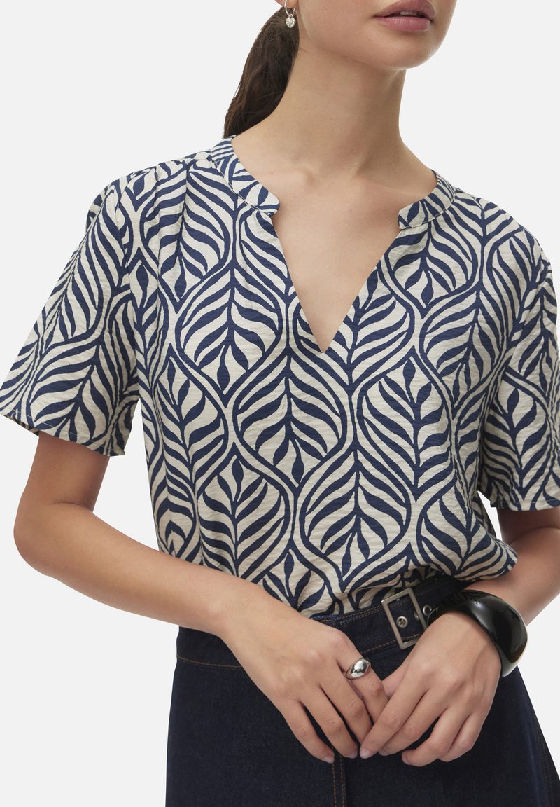 Mujer con blusa azul marino y blanca con estampado de hojas y escote en V, pantalones oscuros con cinturón, anillo de plata, pulsera negra y pendientes en forma de corazón.