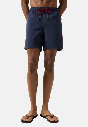 Männliches Model trägt marineblaue Badeshorts mit roten Seitenstreifen und rotem Kordelzug, kombiniert mit schwarzen Flip-Flops vor einem schlichten Hintergrund.