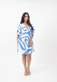 Ein blau-weiß gemustertes Kleid mit tiefem V-Ausschnitt, kurzen, schwingenden Ärmeln und lockerer Passform, getragen mit klaren Absatzsandalen.
