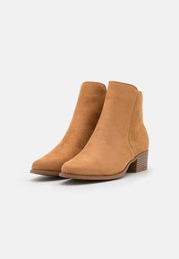 Beżowe zamszowe botki do kostki z ostrym noskiem i niskim, układanym obcasem. Materiał jest miękki, z minimalną ilością szwów i bez widocznych elementów metalowych.