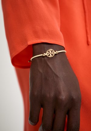 Gullfarget armbånd med en oval anheng og mønstrete detaljer, med en hvit stropp, båret på en mørkebrun hånd. Lys oransje skjorte i bakgrunnen.