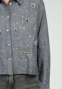 Chemise en lin gris au design court, ornée d'embellissements en forme de fleurs variées et de sequins décoratifs argentés et bleus.