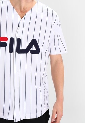 Chemise blanche à boutons avec des rayures verticales bleu marine, manches courtes, et un logo FILA rouge et bleu bien visible à l'avant.