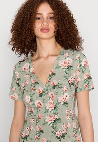 Vestido floral em tecido verde, com decote em V e mangas curtas. O padrão consiste em rosas pêssego e brancas com folhas verdes.