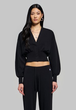 Jeune femme aux longs cheveux noirs porte un cardigan court noir boutonné et un pantalon assorti, accessoirisés avec des boucles d’oreilles créoles en argent et des colliers.