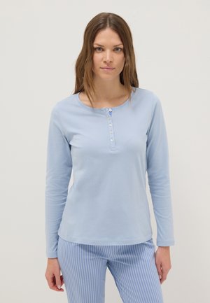Vrouw draagt een lichtblauw Henley-shirt met lange mouwen en blauw-wit gestreepte broek, staand tegen een effen lichte achtergrond.