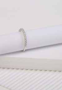 Silberring mit schmalem Band, besetzt mit kleinen klaren Steinen entlang der oberen Kante, platziert auf einer glatten weißen zylindrischen Oberfläche.