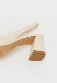 Beige slingback-hæl med firkantet blokhæle, glat læderoverdel og et minimalistisk design. Gyldne hardware-detajler nær hælen.