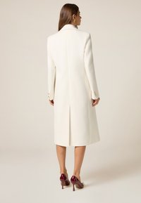Witte op maat gemaakte jas met een enkele split, lange mouwen en knoopmanchetten, gecombineerd met bordeauxrode hoge hakken en minimale accessoires.