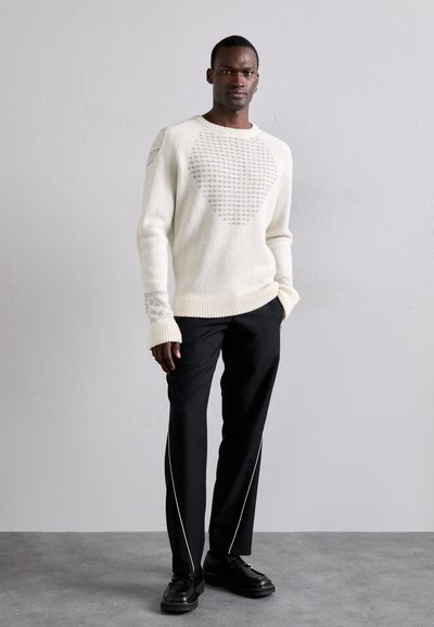Strickpullover in Elfenbein mit strukturiertem Muster, gerippten Bündchen und Rundhalsausschnitt, kombiniert mit schwarzen Hosen mit weißen Seitenstreifen und schwarzen Schuhen.