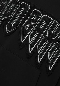Schwarze Kapuzenjacke mit großen, weißen, gestickten Texten mit kantigen Kanten und strukturierten Details. Das Material ist weich und hat eine glatte Oberfläche.