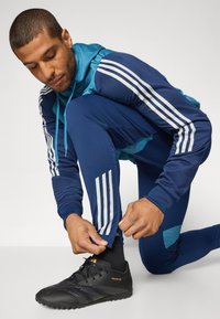 Marineblå atletiske joggingsbukser med hvide striber på ærmerne, blå accenter og sorte fodboldstøvler med struktureret overflade. Tætsiddende design.