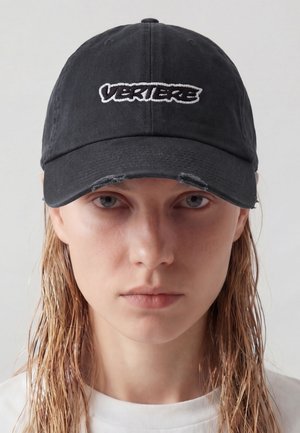 Joven mujer con cabello rubio mojado, lleva una gorra negra con texto blanco "VERIERE" y una camiseta blanca, frente a un fondo claro y liso.