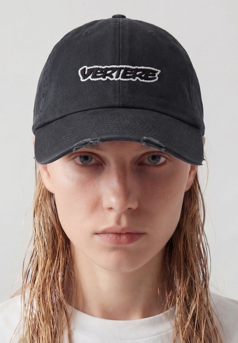 Giovane donna con capelli biondi bagnati che indossa un berretto nero con la scritta bianca "VERIERE" e una maglietta bianca, su uno sfondo chiaro uniforme.