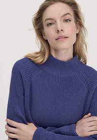 Maglione viola lavorato a maglia con collo alto e maniche raglan, caratterizzato da un tessuto testurizzato e una vestibilità ampia. Braccia incrociate davanti.