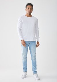 Fehér, hosszú ujjú póló kerek nyakkivágással, világoskék, slim-fit farmernadrággal és fehér sportcipővel egy világos háttér előtt.