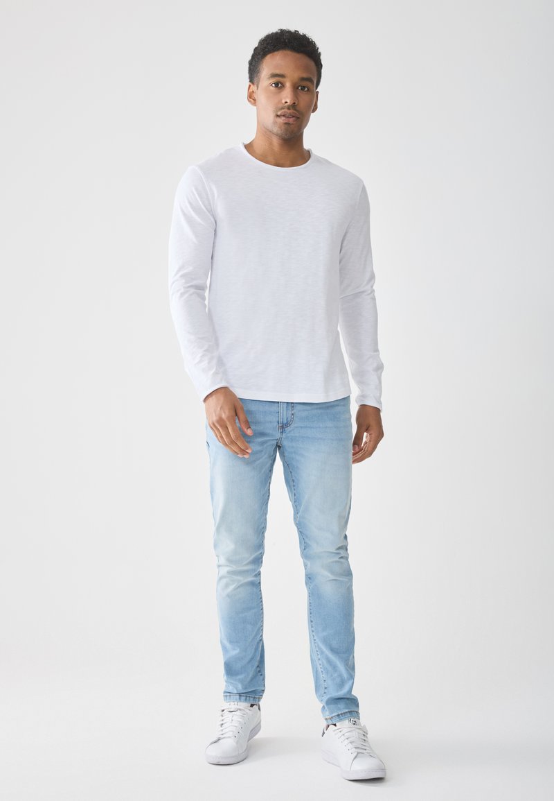 Biała koszula z długim rękawem i okrągłym dekoltem, w zestawieniu z jasnoniebieskimi dopasowanymi dżinsami i białymi trampkami na jasnym tle.