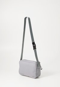 Borsa a tracolla grigia chiara, con forma rettangolare, tessuto morbido, chiusura con zip e una tracolla grigia regolabile con fibbia in plastica.