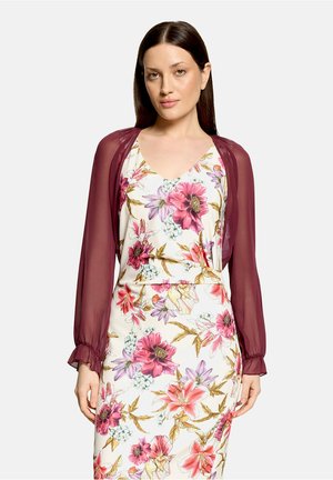 Femme portant une robe blanche avec des motifs floraux roses et violets et un shrug long transparent bordeaux, debout devant un fond uni.