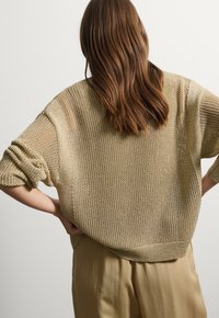 Maglione oversized beige a maglia con maniche ampie, caratterizzato da un motivo testurizzato. Indossato con pantaloni beige chiaro e posizionato su uno sfondo neutro.