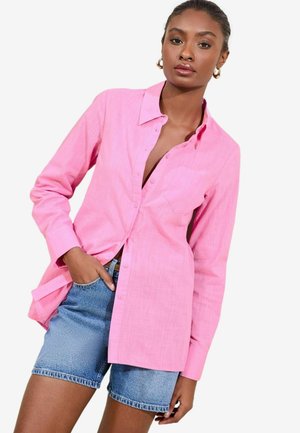 Camicia rosa a maniche lunghe con chiusura a bottoni e taschino sul petto, abbinata a pantaloni corti in denim blu. La camicia presenta un tessuto leggero e strutturato.