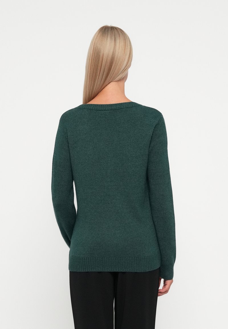 Femme aux longs cheveux blonds portant un pull en maille vert foncé et un pantalon noir, vue de dos sur un fond uni.