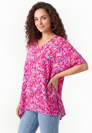 Vrouw met krullend haar die een losse roze top met patroon en een blauwe spijkerbroek draagt, glimlachend met één hand op haar heup tegen een witte achtergrond.