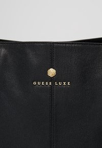 Sort læderhåndtaske med en tekstureret overflade, guldfarvet logo "GUESS LUXE" og en blomsterdetalje i midten.
