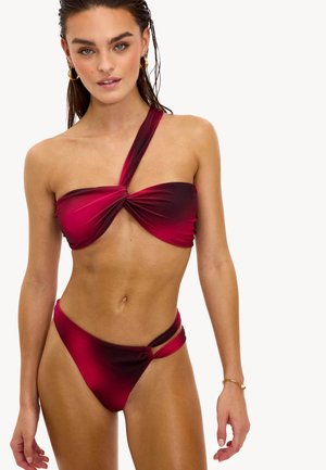 Femme portant un bikini asymétrique dégradé rouge et noir, avec des boucles d'oreilles anneau dorées et un bracelet doré au poignet droit.