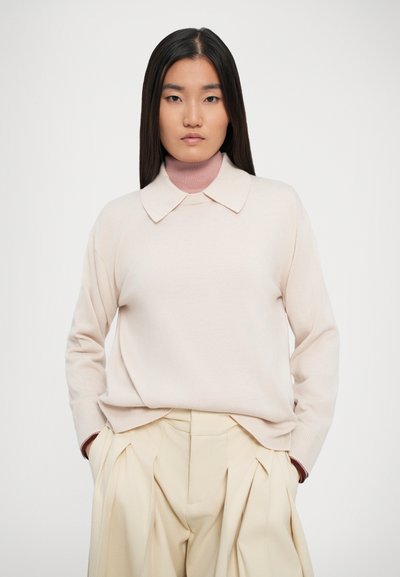Maglione a maglia rosa chiaro con colletto, abbinato a pantaloni beige a vita alta plissettati, dotati di maniche lunghe e una vestibilità ampia.