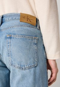 Jeans de mezclilla azul claro con un parche de cuero marrón de Calvin Klein Jeans en la cintura. Presentan bolsillos traseros clásicos y detalles de costura visibles.