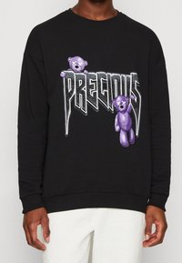 Felpa nera con il testo "PRECIOUS" in stile screpolato e due orsetti viola con gli occhi sbarrati sulla parte anteriore, indossata con pantaloni bianchi.