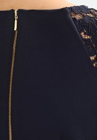 Navy dress featuring a gold zipper closure at the back, with intricate black lace detailing on the shoulder area and smooth fabric texture.