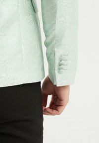 Veste de couleur vert clair à motif paisley avec un tissu texturé ; présente des poignets boutonnés et une coupe ajustée, montrée de dos et de côté.