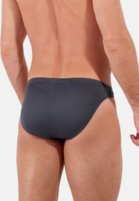 Dunkelgraue Schwimmslips mit glatter Textur, niedrig sitzendem Design und minimaler Rückendeckung. Verfügt über einfache Nähte entlang der Ränder.