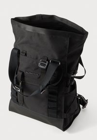 Zaino a sacca nera realizzato in tela resistente, con spallacci regolabili, tasche laterali e una base liscia e testurizzata. Presenta dettagli in metallo.