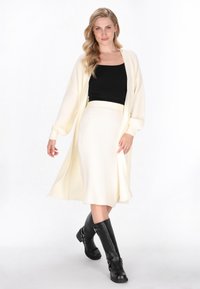 Langer, gerippter Cardigan in Cremeweiß, kombiniert mit einem passenden gerippten Rock. Schwarzes enges Oberteil und knielange schwarze Lederstiefel.